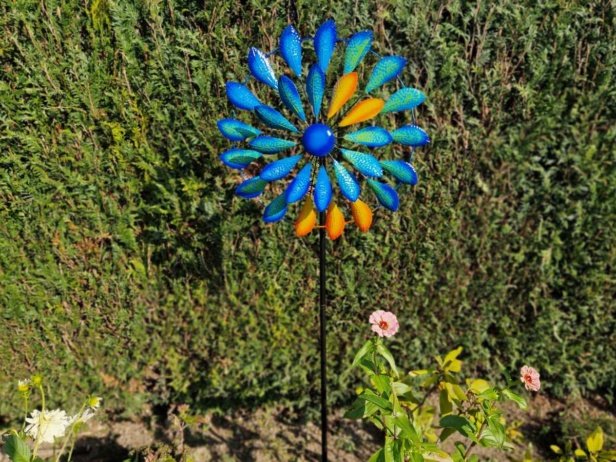 Buntes Gartenwindrad aus Metall in Blau, Gelb und Grün auf einem Stab im Garten.